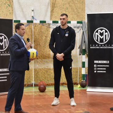 Trzech mężczyzn stoi na sali gimnastycznej. Jedn z garniturze trzyma piłke do siatkówki oraz mikrofon do którego przemawia.