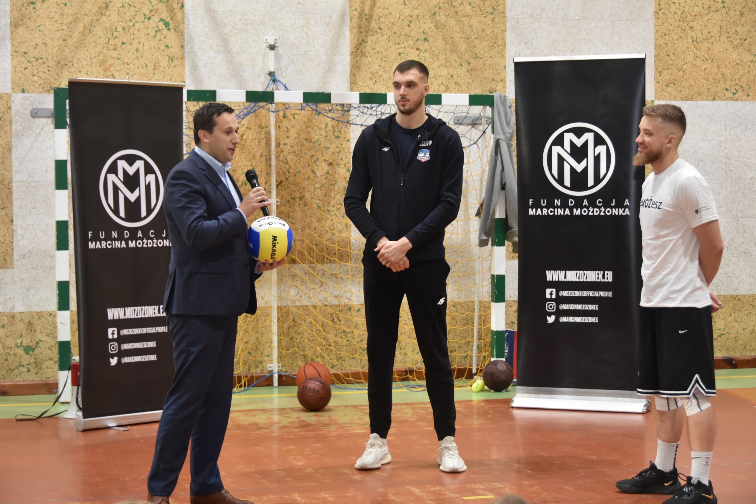 Trzech mężczyzn stoi na sali gimnastycznej. Jedn z garniturze trzyma piłke do siatkówki oraz mikrofon do którego przemawia.