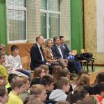 Grupa dzieci i gości trzech mężczyzn w garniturach i kobieta o blond włosach ubrana elegancko w marynarkę brązową siedzą na sali gimastycznej w szkole.