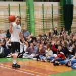 Na sali guimnastycznej siedzi grupa dzieci, przed nimi mężczyzn stoi ubrano na sportowo i kręci piłką do koszykówki.