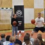 Młody m,ężczyzna ubrany na sportowo trzyma w rękach piłki do koszykówki, obok stoi dziewczynka też trzyma piłkę do koszykówki. Obydwoje są na sali gimnastycznej. Przed nimi widownia składająca się z dużej grupy dzieci.