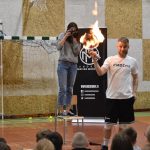 Młody mężczyzna trzyma zapaloną ogniem piłką, znajduje się na sali gimnastycznej. Przed nim widownia składająca się z dzieci.