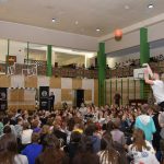 Młody chłopak w tłumie dzieci na sali gimnastycznej rzuca piłką do kosza.