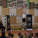 Młody chłopak ubrany na sportowo wspina się na kosza do koszykówki na sali gimnastycznej. Jgo poczyniania obserwuje grupa dzieci.