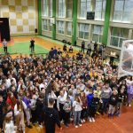 Grupa dzieci i nauczycieli stoi na sali gimnastycznej pod koszem do koszykówki, i słuchają młodego mężczyzny który stoi na tymk koszu.