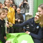 Młoda dziewczynka o jasnych włosach robi sobie zdjęcie z młodym mężczyzną telefonym komórkowym. Przed nim stolik i grupka dzieci.