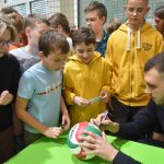 Młody meżczyzna podpisuje się na piłce do siatkówki, obsewuje to grupa dzieci.