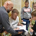 Młody mężczyzna wpisuje się do zeszytuna sali gimnastycznej. Zeszyt trzyma starszy łysy mężczyzna. Przed nimi stoi grupa dzieci.