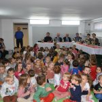 Grupa dzieci siedzi na sali przedszkolnej, za nimi goście ubrani w odświętne stroje siedzą na stołem nakrytym białem obrusem.