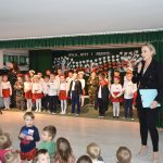 Kobieta o blond włosach ubrana w elegancki gartiur czarny stoiw sali szkolnej i przemawia do dzieci. Za nią w odświętnych strojach stoją dzieci.