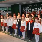 Grupa dzieci w odświętnych strojach stoi w sali i występuje przed publicznością.