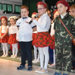 Trójka dzieci, dziewczynka w środku ubrana w spódniczkę i biała koszulę oraz dwóch chłopaków mówi do mikrofonu wiersz, za nimi reszta dzieci.