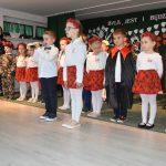 Grupa dzieci, dziewczynek i chłopców w odświętnych strojach mówi wierszyki do mikrofonu na sali szkolenj.