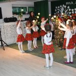 Grupa dziewczynek ubranych w białe bluzki i czerowne spódniczki trzyma białe róże wysoko, a obok nich przechodza chłopcy ubrani w stroje żołnierskie.