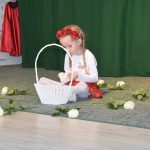 Dziewczynka w białej koszulce rwie białe róże do koszyka białego. Siedzi w sali przedszkolnej na dywanie. Za nią zielona kurtyna.