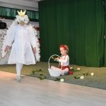 Dziewczynka przebrana w strój orła białego krąży koło drugiej dziewczynki, kóra siedzi na podłodze i rwie białe róże do koszyka.