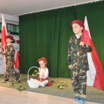 Trójka dzieci, dwóch chłopców ubranych w żołnierskie stroje trzymają w rękach duże flagi biało czerowne. Na podłodze siedzi dziewczynka w białej koszulce i rwie białe róże do koszyka.