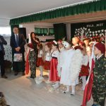 Grupa dzieci ubrana w odświętne stroje słucha dwóch mężczyzn w garntiurach, którrzy przemawiają do mikrofonu.