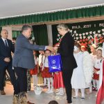 Starszy mężczyzna w garniturze przekazuje na ręce młodej kobiety o blond włosach i czarnym garniturze granatową torbę prezentową z herbem gminy. Za nimi stoją dzieci w odświętnych strojach.