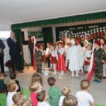 Grupa dzieci przebrana w odświętne stroje stoi na sali gimnastycznej i słucha jak przemawiają do nich goście ( dwóch meżczyzn w garniturach).