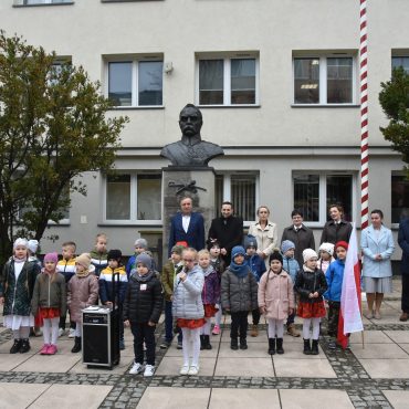 Grupa kilkudziesięciu osób (dzieci oraz kobiet i mężczyzn) stoją na kostce brukowej szarej przed jasnym budynkiem. Za nimi pomnik przedstawiające półpiersie Józefa Piłsdudskiego.