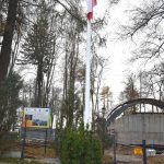 Masz flagowy z biało-czerwoną flagą. Z tyłu widać drzewa i krzewy oraz plac budowy. Pogoda jest zachmurzona.