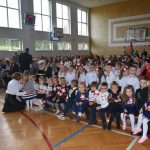 Duża grupa dzieci i osób dorosłych siedzących na rozległej sali gimnastycznej. Większość osób jest ubrana w odświętne stroje.