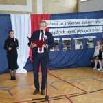 Mężczyzna w sile wieku przemawia za pomocą mikrofonu patrząc w notatki. Stoi na rozległej sali gimnastycznej. Za nim widać grupę elegancko ubranych dziewcząt