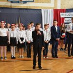 Grupa elegancko ubranych dzieci stojących przed ozdobną zasłoną. Jeden chłopak ubrany w czarny garnitur trzyma mikrofon i wykonuje występ muzyczny.