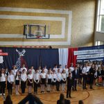 Grupa elegancko ubranych dzieci stojących przed ozdobną zasłoną. Jeden chłopak ubrany w czarny garnitur trzyma mikrofon i wykonuje występ muzyczny.