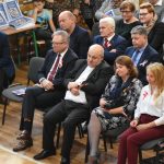 Duża grupa osób obojga płci siedząca na krzesłach na rozległej sali gimnastycznej. Uważnie spoglądają przed siebie.