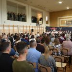 Duża grupa dorosłych osób siedzących na prostych krzesłach w rozległej sali gimnastycznej. Z porzodu widzimy elegancko ubraną grupę dzieci w trakcie występu artystycznego.