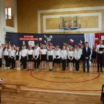 Duża grupa osób obojga płci pozująca do zdjęcia na sali gimnastycznej. Wszyscy są odświętnie ubrani. Część z zebranych osób macha flagami.