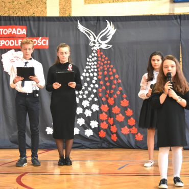 Cztery młode osoby (trzy dziewczyny i chłopak) stojący w odświętnych strojach na sali gimnastycznej. Z tyłu widać szara kurtynę z napisem 11 Listopada Święto Niepodległości