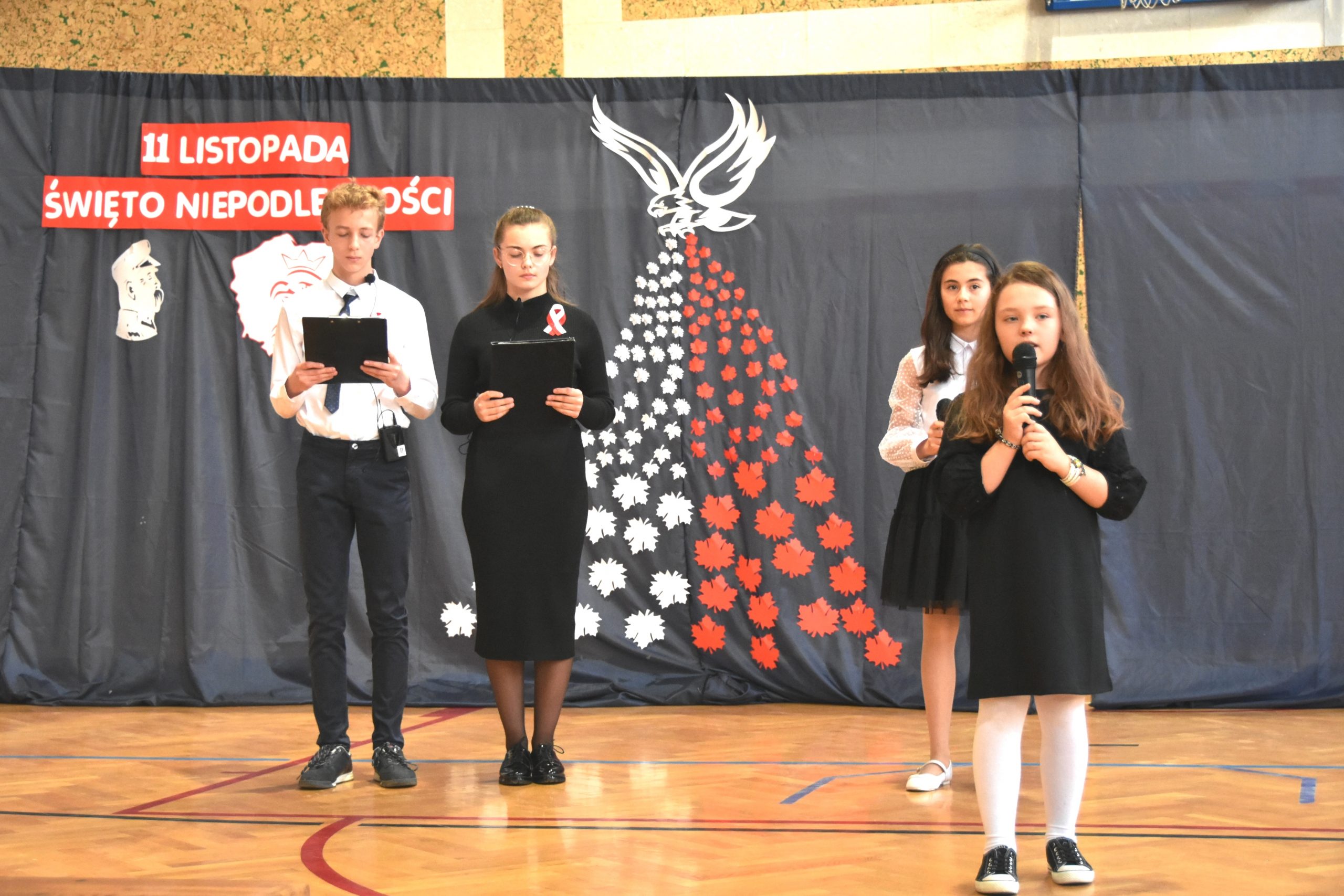 Cztery młode osoby (trzy dziewczyny i chłopak) stojący w odświętnych strojach na sali gimnastycznej. Z tyłu widać szara kurtynę z napisem 11 Listopada Święto Niepodległości