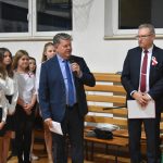 Dwóch dorosłych mężczyzn w garniturach stojących na sali gimnastycznej. Jeden z nich przemawia za pomoca mikrofonu drugi trzyma notatki. Za nimi widać grupę dzieci.