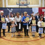 Grupa osób w odświętnych strojach pozująca do zdjęcia na rozległej sali gimnastycznej. Głownie są to dzieci i młodzież oraz dwie starsze panie oraz starszy mężczyzna.