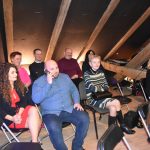 Grupa osób różnej płci i wieku siedząca na rozległym drewnianym poddaszu.