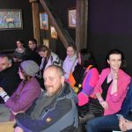 Grupa osób dorosłych różnej płci i wieku siedząca na rozległym drewnianym poddaszu i patrząca w przód.