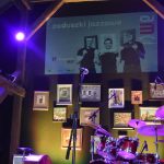 Drewniana scena na której znajdują się perkusja. Z tyłu na ścianie widać obraz wyświetlacza z napisem Zaduszki Jazzowe..