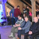 Grupa osób dorosłych różnej płci i wieku siedząca na rozległym drewnianym poddaszu.