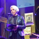 Kobieta ubrana wieczorowo z białymi włosami oraz białymi koralami. Przemawia za pomocą mikrofonu spoglądając przed siebie.