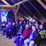 Grupa osób dorosłych różnej płci i wieku siedząca na rozległym drewnianym poddaszu i patrząca w przód.