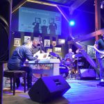 Grupa muzyczna złożona z czterech mężczyzn grajaca na instrumentach muzyczny na drewnianej scenie w rozległym pomieszczeniu.