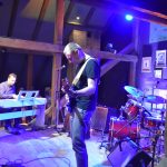 Grupa muzyczna złożona z czterech mężczyzn grajaca na instrumentach muzyczny na drewnianej scenie w rozległym pomieszczeniu.