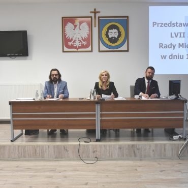 Trzy osoby (dwóch dorosłych mężczyzn i kobieta zasiadający przy biurku). Na ścianie widać herby i obraz wyświetlacza.