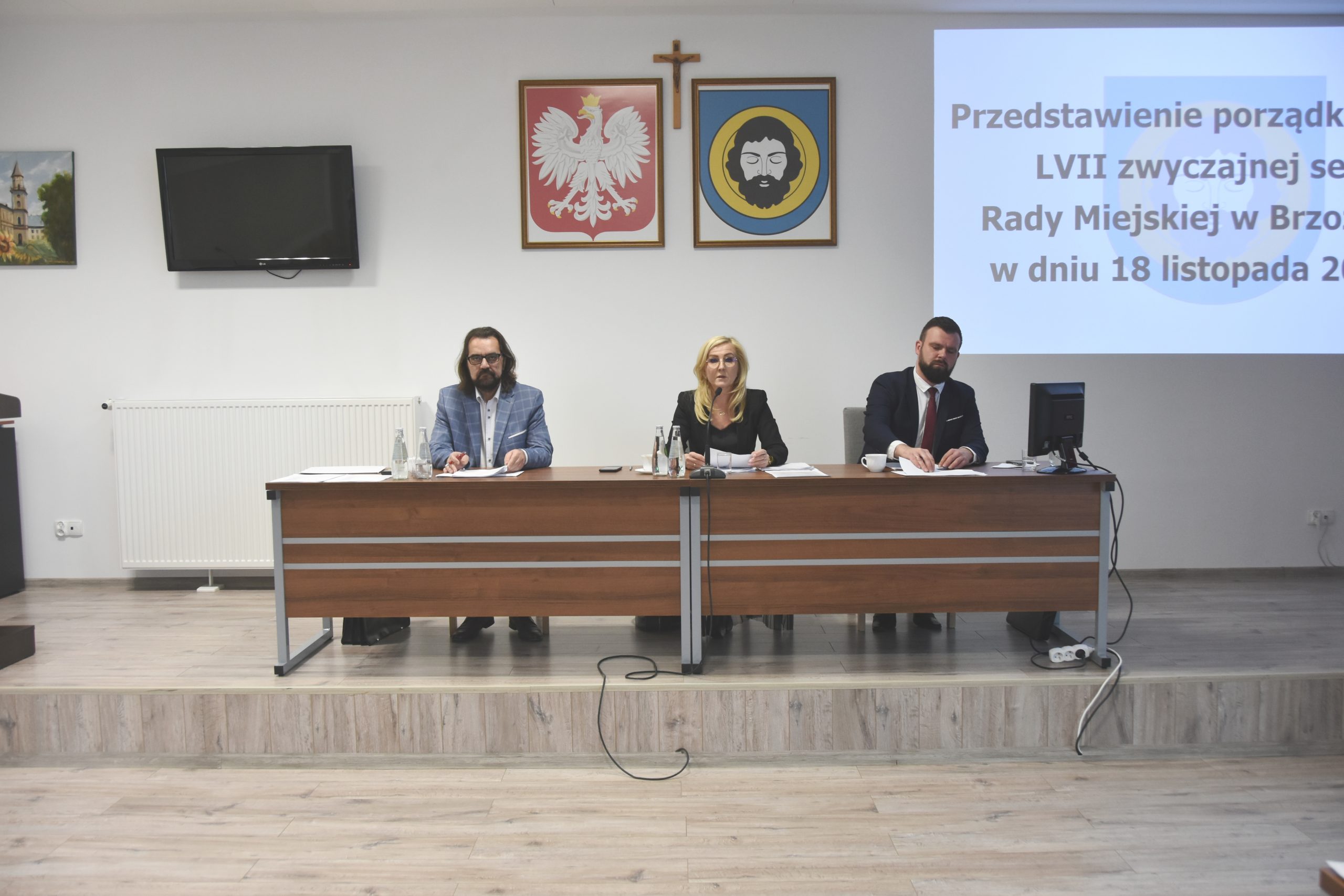 Trzy osoby (dwóch dorosłych mężczyzn i kobieta zasiadający przy biurku). Na ścianie widać herby i obraz wyświetlacza.