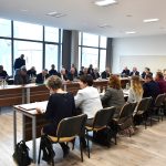 Grupa osób różnej płci i wieku zasiadający przy biurkach. Na ich blatach widać kartki papieru oraz wodę. Wszyscy znajdują się na rozległej sali.