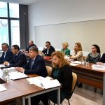 Grupa osób różnej płci i wieku zasiadający przy biurkach. Na ich blatach widać kartki papieru oraz wodę. Wszyscy znajdują się na rozległej sali.