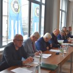 Grupa dorosłych mężczyzn zasiadających przy biurkach. Na ich blatach widać kartki papieru oraz wodę. Wszyscy znajdują się na rozległej sali.